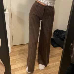Bruna randiga byxor från H&M Divided - Snygga bruna byxor med vita ränder från H&M Divided. De har en högmidjad passform och raka ben som ger en stilren look. Jag är 160cm och har vanligtvis 34 i storlek