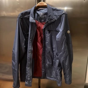 Mörkblå jacka från Snoot - Snygg mörkblå jacka från Snoot med dragkedja och flera praktiska fickor. Jackan har en stilren design med långa ärmar och en diskret logga på ärmen. Perfekt för höst och vår.