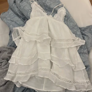 Vit klänning från Gina Tricot - Söt vit klänning från Gina Tricot med volanger och spetsdetaljer. Klänningen har smala axelband och en luftig design, perfekt för varma dagar. Den har en romantisk känsla med sina lager av volanger och spets.