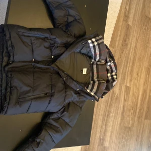 Svart pufferjacka med rutigt foder - Säljer en svart pufferjacka från Burberry med ett snyggt rutigt foder i huvan. Jackan har dragkedja och knappar framtill samt långa ärmar. Perfekt för kyliga dagar. (Priset kan diskuteras)