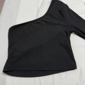 Svart ribbad oneshoulder topp - Snygg svart ribbad topp med oneshoulder-design och lång ärm. Perfekt för en stilren look. Öppen i ryggen. 