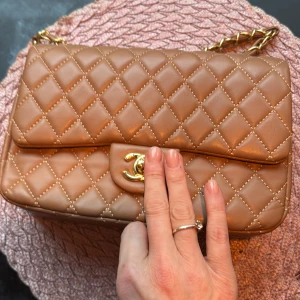 Beige quiltad axelväska med guldkedja - Snygg beige axelväska med quiltat mönster och guldkedja. Väskan har en klassisk design med en elegant guldfärgad logotyp på framsidan. Insidan är klädd i rött tyg, vilket ger en fin kontrast. Perfekt för att ge din outfit en lyxig touch. På sista bilden syns de att lädret hoppat ur öglan.