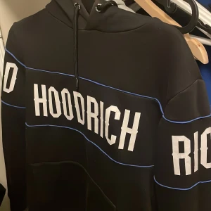 Svart hoodie från Hoodrich - Säljer en svart hoodie från Hoodrich med vit text och blå detaljer. Den har en klassisk design med huva och dragsko. Perfekt för en avslappnad stil.