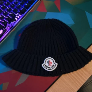 Svart ribbad mössa från Moncler - Snygg svart ribbad mössa från Moncler med ett broderat märke framtill. Perfekt för att hålla värmen under kyliga dagar.