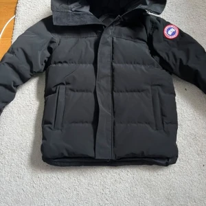 Svart dunjacka från Canada Goose - Säljer en svart dunjacka från Canada Goose med deras ikoniska märke på ärmen. Jackan har en dragkedja framtill och är perfekt för kalla vinterdagar. Den är långärmad och har en stilren design som håller dig varm och bekväm.