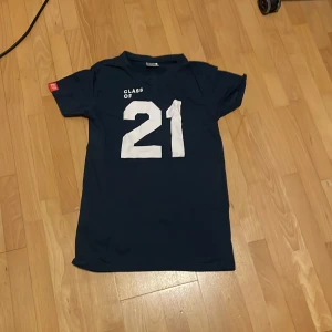 Mörkblå t-shirt med tryck 'Class of 21' - Säljer en mörkblå t-shirt från EF med stort vitt tryck 'Class of 21' på framsidan. T-shirten är kortärmad och tillverkad i 100% bomull, vilket gör den mjuk och bekväm att bära. Perfekt för en avslappnad stil.
