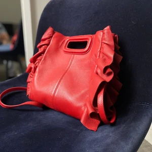 Röd axelremsväska Maje - En klassisk klarröd axelremsväska från Maje. Väskan köptes in 2019, är använd men i gott skick. Till väskan kommer ett justerbart axelremsband som också går att byta ut mot andra band eller kedjor. Väskan är helt läder och har måtten 21x20x6 exklusive volangerna på sidan.
