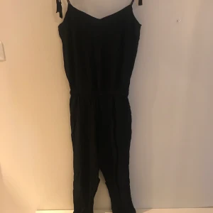 Jumpsuit svart i stl M - Svart jumpsuit med knytbara axelband, oanvänd, brodyr på bröstet, lite resår i midjan 