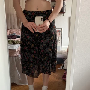 Svart blommig långkjol - Svart kjol med vita prickar och färgglada blommor. Står storlek XXL men funkar bra att sätta fast med säkerhetsnålar om man vill ha mindre (andra bilden är hur den sitter på mig med storlek s utan nålar). Möts i Stockholm annars betalar köpare frakt. 