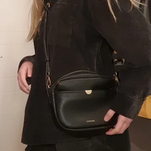 Svart dondonna väska - Jättefin svart dondonna väska i fake skinn med guldiga detaljer. 😇 Det går att både spänna och ta lös bandet. Den är inte så mycket använd den har bara varit hängd på min krok i ca 1,5 år.😙 Den ser Väldigt ny ut!!😋jag köpte den för 600 kr i butik. FRAKT: 66kr så sammanlagt blir det 216 kr.