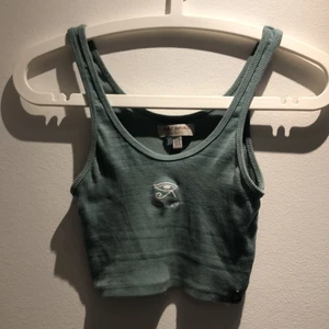 Grön topp storlek XS - Det är en fin grön croppad topp med ett vitt öga i mitten som detalj. Jag köpte den för ca ett år sen men har knappt använts så den är i väldigt gott skick.      Jag köpte den second hand här på plick! 