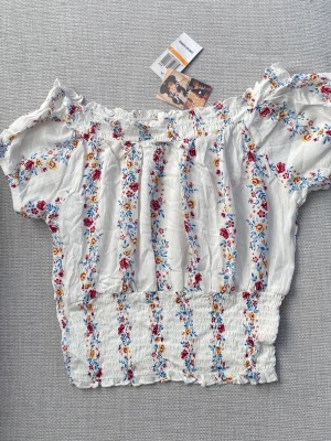 Ny blommig offshoulder topp från SELF♥️E - Supersöt och somrig offshoulder topp från SELF♥️E i vitt med blommigt mönster i rött, blått och gult. Toppen har korta puffärmar och smockad midja. Ny och oanvänd med prislappen kvar. Storlek: S