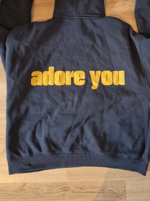 Marinblå hoodie med gul text - Cool blå hoodie med huva och ficka framtill. På ryggen står det 'adore you' i stora, gula bokstäver. Hoodien har ribbade muddar vid ärmar och nederkant. Perfekt för dig som gillar statement-plagg.