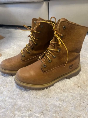 Bruna Timberland kängor i mocka - Säljer ett par klassiska bruna Timberland kängor i mocka med högt skaft och robust sula. Kängorna har gula snören och metallöljetter, samt Timberland-loggan på sidan. Perfekta för dig som vill ha en stilren och tålig sko till hösten.