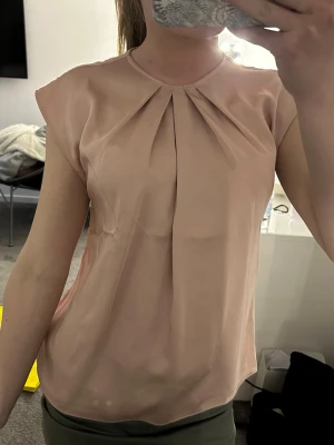 Ljusrosa blus med veckad front - Snygg ljusrosa blus i ett glansigt, mjukt material med veckade detaljer framtill vid halsen. Blusen har en rak passform och korta ärmar som ger en avslappnad men ändå chic look. Perfekt att styla med både jeans och kjol.