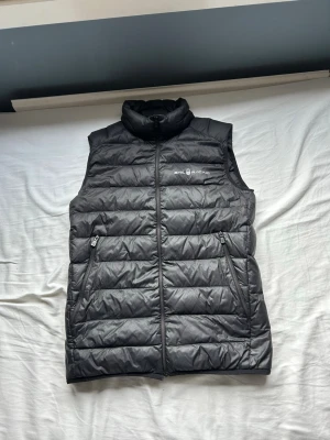 Svart dunväst från Sail Racing - Snygg svart dunväst från Sail Racing. Perfekt för lager-på-lager och håller dig varm under kyliga dagar. Den har två stora fickor. Samt lite slitet vid början av dragkedjan men kan fixas. Annars inga defekter alls! Nypris 1200. Hör av er vid funderingar✌🏼