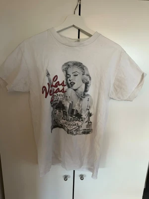 Vit Las Vegas t-shirt med tryck - Säljer en vit t-shirt med coolt Las Vegas-tryck framtill i svart, grått och rött. Motivet har en retrovibe med stadssilhuett och text. Klassisk passform och rund hals. Perfekt för dig som gillar statement-plagg och streetstyle. Köpt lokalt i Las Vegas. Storlek M