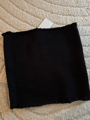 Svart smockad tubtopp - Säljer en svart smockad tubtopp med stretchig passform och små volangdetaljer upptill och nedtill. Perfekt att styla till jeans eller kjol för en trendig look. Enkel och stilren design som passar till mycket.