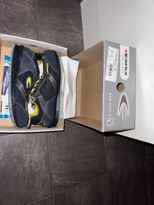 Svarta och gula sneakers från Cofra - Snygga sneakers från Cofra i svart med gula detaljer och grå sula. Skorna har en sportig design med meshpaneler och förstärkta partier. Perfekta för dig som gillar en cool och modern look. Skosnören i svart och gult ger extra edge. Arbetsskor helt nya