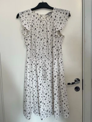 Vit blommig klänning med volang - Söt vit klänning med små svarta blommor, volangdetaljer vid axlarna och knytband i midjan. Klänningen har en luftig passform och är ärmlös med rund halsringning. Perfekt för varma dagar och ger en romantisk vibe.