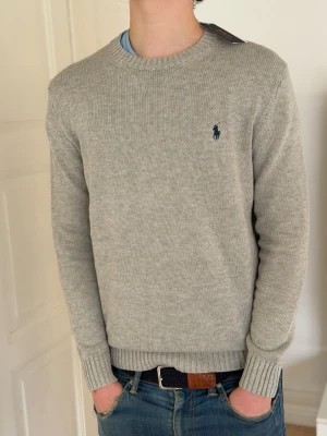 (Helt ny)Ljusgrå stickad tröja från Polo Ralph Lauren - (Moddelen är 187 och väger 77 kg )Snygg ljusgrå stickad tröja från Polo Ralph Lauren med rund halsringning och diskret blå logga broderad på bröstet. Tröjan har ribbade muddar vid ärmslut och nederkant, perfekt för lager på lager och enkel att matcha.