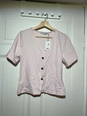Ljusrosa v-ringad topp med knappar - Säljer en ljusrosa topp från 157 med v-ringning och tre svarta knappar framtill. Toppen har korta ärmar och en enkel, stilren design som passar perfekt till jeans eller kjol. Materialet känns mjukt och lätt, perfekt för varmare dagar.