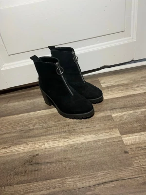 Svarta stövletter med grov klack - Snygga svarta stövletter i mockaimitation med chunky klack och grov sula. Dragkedja framtill med rund metallring som detalj. Skorna har en modern siluett och passar perfekt till jeans eller kjol. Märke ej synligt på bilderna.