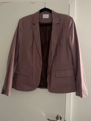 Brun kavaj - Stilren brun kavaj med två knappar framtill. Kavajen har en rak passform. En knapp vid ärmen är lite skadad, se bild 4, i övrigt väldigt bra skick. 