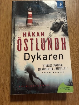 Dykaren av Håkan Östlundh pocket - Spännande kriminalroman i pocketformat av Håkan Östlundh. Omslaget är färgstarkt med en fyr, nycklar och en dykare mot en dramatisk bakgrund. pocketbok!  Perfekt för dig som gillar deckare. Det är andra boken i Östlundhs serie om kriminalpolisen Fredrik Broman