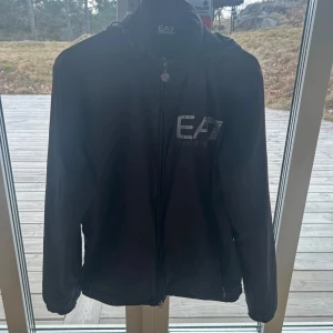 Svart jacka från Emporio Armani EA7 - Snygg svart jacka från Emporio Armani EA7 med dragkedja och huva. Perfekt för en stilren look. Jackan har en diskret logga på bröstet och är tillverkad i ett syntetmaterial. Super fint skick. 