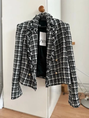 Rutig svartvit kavaj från Zara - Stilren svartvit rutig kavaj från Zara med guldknappar och fransade kanter. Kavajen har en klassisk passform och är tillverkad i ett strukturerat tyg som ger en exklusiv känsla. Perfekt för dig som vill ha en trendig och elegant look.