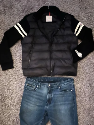 Svart dunväst från Moncler - Säljer en svart dunväst från Moncler med quiltad design och dragkedja framtill. Västen har två sidofickor med dragkedja och ribbstickade detaljer på kragen. Klassisk Moncler-logga på ärmen och vita ränder på axlarna ger en sportig touch.