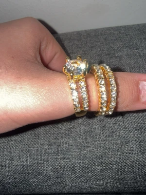 Guldfärgade ringar med stenar - Två guldfärgade ringar med gnistrande klara stenar runt hela banden. Den ena ringen har en stor rund sten i mitten som verkligen fångar blicken. Båda ringarna har dubbla rader av små stenar för extra bling. Perfekt för dig som gillar statement-smycken.