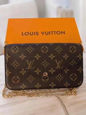 Iouis Vuitton handväska  - Butikspriset är 16 000kr.Väska/Kuvertväska: En rektangulär väska med ett spetsigt lock (envelop-stil) som stängs med en guldfärgad tryckknapp. Den är klädd i ett mörkt brunt mönster med geometriska former och blomliknande symboler. Blixtlåsfodral: Ett platt fodral i samma mönstrade canvas som väskan, försett med en mörk dragkedja längs ovansidan och en liten dragkläpp i läder.