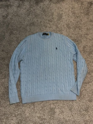 Ljusblå kabelstickad tröja Polo Ralph Lauren - Snygg ljusblå kabelstickad tröja från Polo Ralph Lauren med klassisk rund halsringning och broderad logga på bröstet. Tröjan har långa ärmar och ribbade muddar vid ärmslut och nederkant. Perfekt för en stilren och avslappnad look.