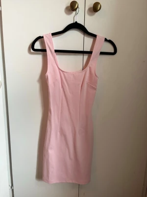 Ljusrosa tight klänning - Säljer en ljusrosa, figurnära klänning med breda axelband och rundad urringning både fram och bak. Klänningen är enkel och stilren, perfekt för dig som gillar minimalistisk design. Aldrig använd.