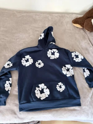 Mörkblå hoodie med vita blommor - Snygg mörkblå hoodie med stora vita blommönster över hela tröjan. Klassisk känguruficka framtill och huva. Perfekt för dig som vill sticka ut med en unik och trendig stil. Mjuk och skön bomullsmix som passar till alla tillfällen. Storlek S men passar M