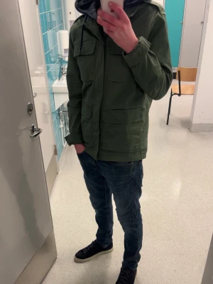 Olivgrön utilityjacka från Weekday - Snygg olivgrön utilityjacka från Weekday med fyra stora fickor framtill, axelklaffar och klassisk krage. Jackan är tillverkad i bomull och har en rak passform med långa ärmar och knappar vid ärmsluten. Perfekt för lager på lager under höst och vår.