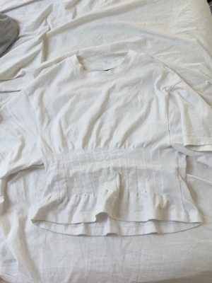 Vit topp med volang från Gina Tricot - Säljer en vit topp från Gina Tricot i bomull med markerad midja och volang nedtill. Toppen har korta ärmar och rund halsringning, samt sydda detaljer som ger en snygg siluett.