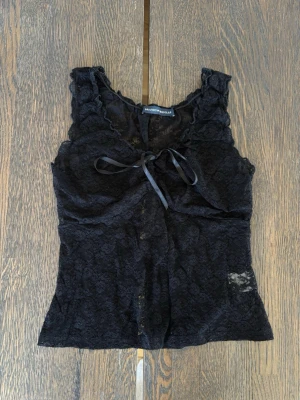 Svart spetslinne från Brandy Melville - Snygg svart ärmlös topp från Brandy Melville i spets med volangkant och söt knytning framtill. Perfekt för dig som gillar en feminin och trendig stil. Tunt och lätt material som känns luftigt.