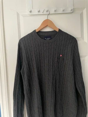 Bondelid Mörkgrå kabelstickad 100% bomull - Snygg mörkgrå kabelstickad tröja från Gant med rund halsringning och diskret Gant-logga i rött och vitt på bröstet. Tröjan har långa ärmar och ribbade muddar vid ärmslut och nederkant. Perfekt för lager på lager och stilren vardagslook. Tröjan är i storleken M och är 100% bomull