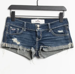 Blå jeansshorts från Hollister - Snygga blå jeansshorts från Hollister med slitna detaljer och fransig kant. Klassisk femficksmodell med låg midja och dubbla knappar framtill. Perfekta för varma dagar och en avslappnad stil.