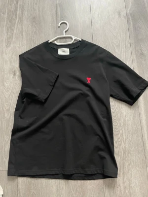 Svart t-shirt från AMI Paris - Svart t-shirt från AMI Paris med klassisk passform och rund hals. På bröstet finns ett litet rött broderat hjärta med A-logga. Tillverkad i mjuk bomull som känns skön mot huden. Perfekt till jeans eller shorts för en clean look.
