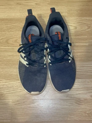 Svarta Adidas sneakers med mesh - Snygga svarta Adidas sneakers med ovandel i mesh och vita detaljer på sidorna. Skorna har svarta snören, orange Adidas-logga på plösen och vit sula. Perfekta för en sportig och avslappnad stil.