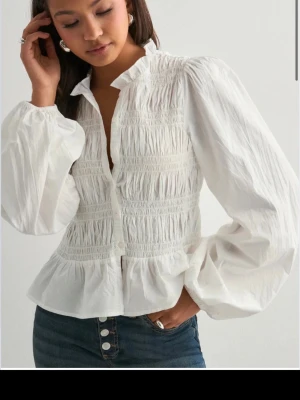 Vit blus med volang och puffärm - Säljer en vit blus med smockdetaljer och volang nedtill. Blusen har puffiga långärmar och en liten volangkrage. Stängs med knappar framtill och är gjord i ett tunt, vävt material som känns luftigt och fräscht.