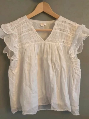 Vit volangblus med spetsdetaljer - Söt vit blus med volangprydda ärmlösa axlar och v-ringning. Blusen har fina spetsdetaljer och lätt rynkade partier upptill. Tillverkad i ett tunt, luftigt tyg som ger en romantisk känsla. Perfekt att styla med jeans eller kjol.