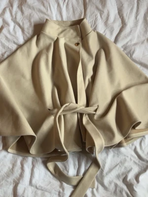 Beige Poncho-kappa - Säljer den snygga beigea poncho-kappan från Nelly💗 One Size, guld detalj och använd 1-2 ggr!! Säljer då jag har en svart som används mer💗