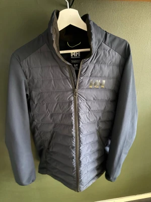 Mörkgrå quiltad pufferjacka Helly Hansen - Snygg mörkgrå pufferjacka från Helly Hansen med quiltad front och rygg, samt hel dragkedja. Jackan har hög krage och två sidofickor. Perfekt för kyliga dagar och har en stilren look med diskret logga på bröstet. Storlek S