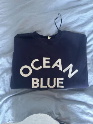 Mörkblå sweatshirt OCEAN BLUE - Mörkblå sweatshirt med stora vita bokstäver där det står 'OCEAN BLUE' på framsidan. Tröjan har rund halsringning och ribbade muddar. Perfekt för en avslappnad och stilren look. Materialet känns mjukt och bekvämt mot huden.
