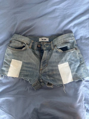 Blå jeansshorts med rå kant från Acne - Säljer ett par blå jeansshorts från Acne med rå, fransig kant och vita patch-detaljer framtill. Klassisk femficksmodell med knappgylf och bälteshällor. Perfekta för en avslappnad och trendig look.